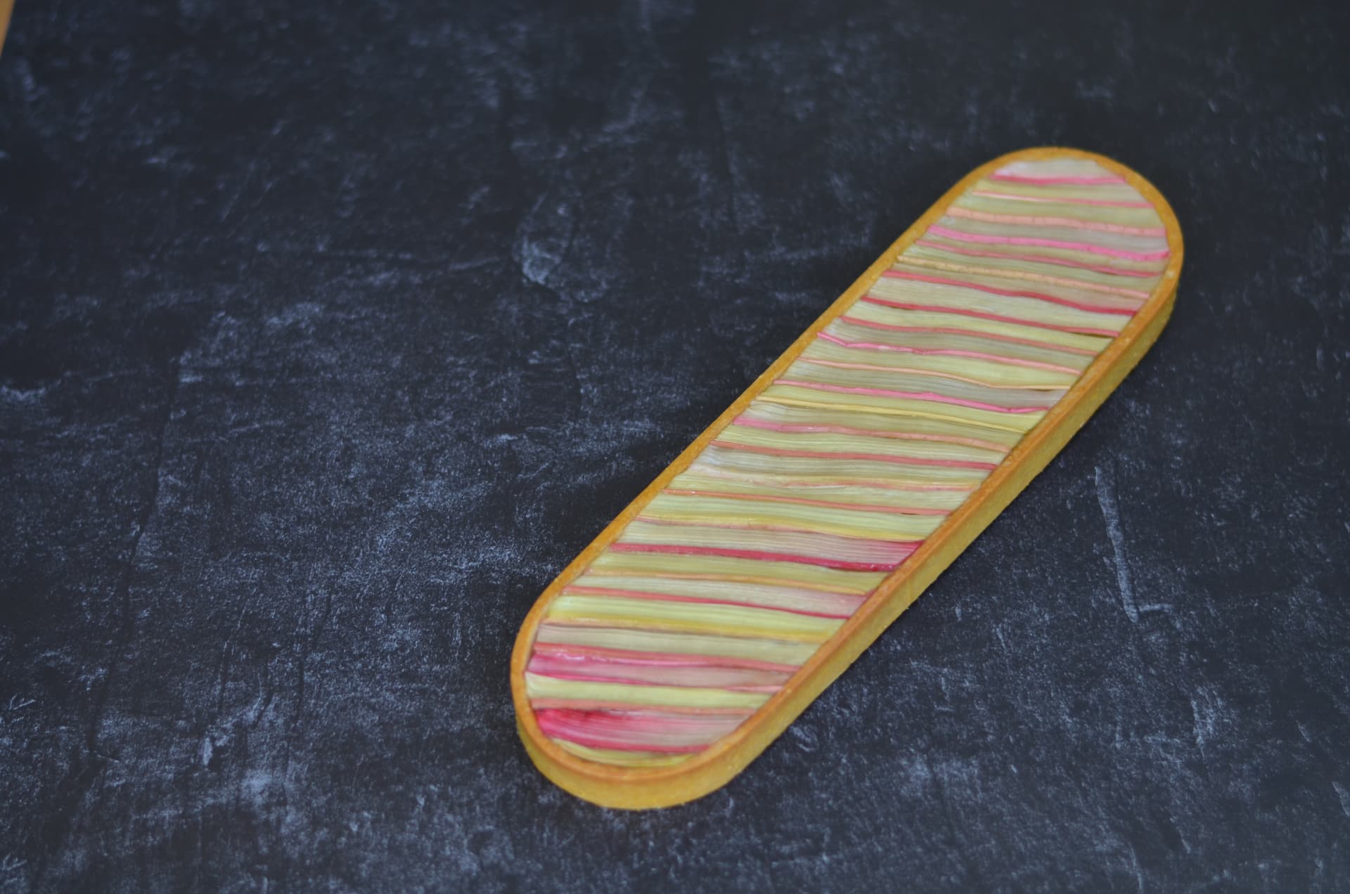 Long rhubarb tart with striped fruit arranged on a dark textured surface